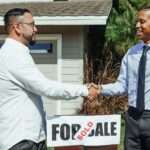 Two men shaking hands in front of house sold sign, sealing real estate deal outdoors.